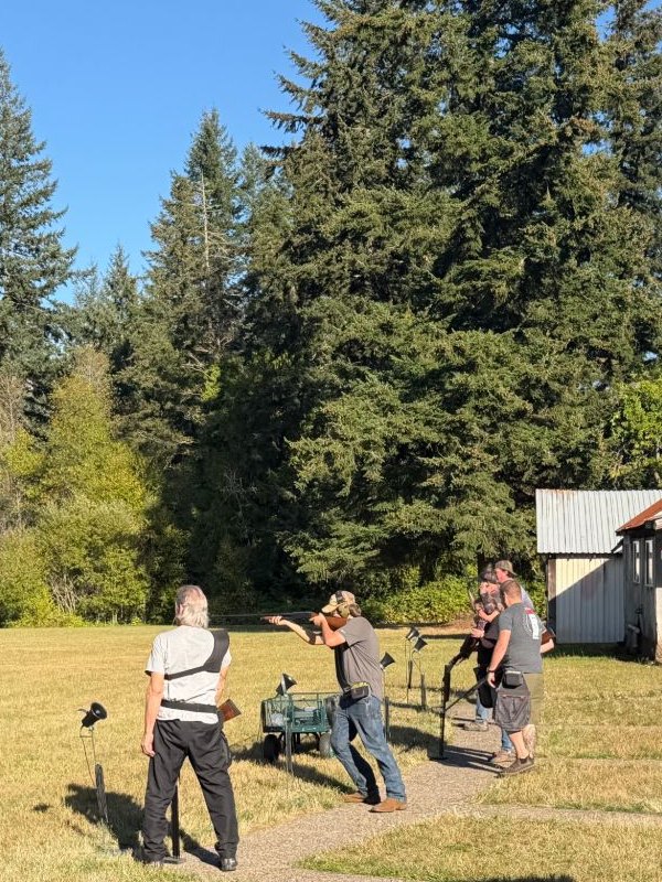 A shooter swings on a target from the trap line on a sunny day, the scorer and the next squad watching, tall firs behind
